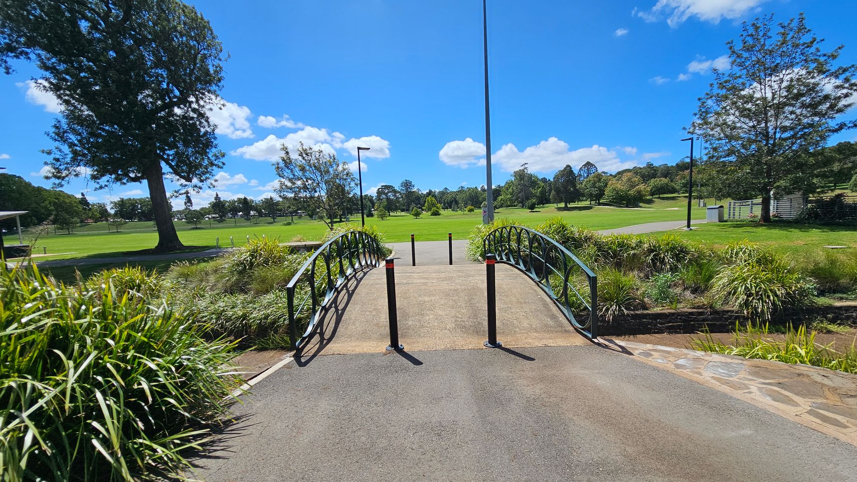 A small arched bridge with black railings crosses over a narrow stream. The bridge is flanked by tall grasses and shrubs. In the background, a vast green park with scattered trees and a few lamp posts stretches out. A large tree stands prominently on the left, while a paved path leads into the distance. The sky is clear with a few clouds.