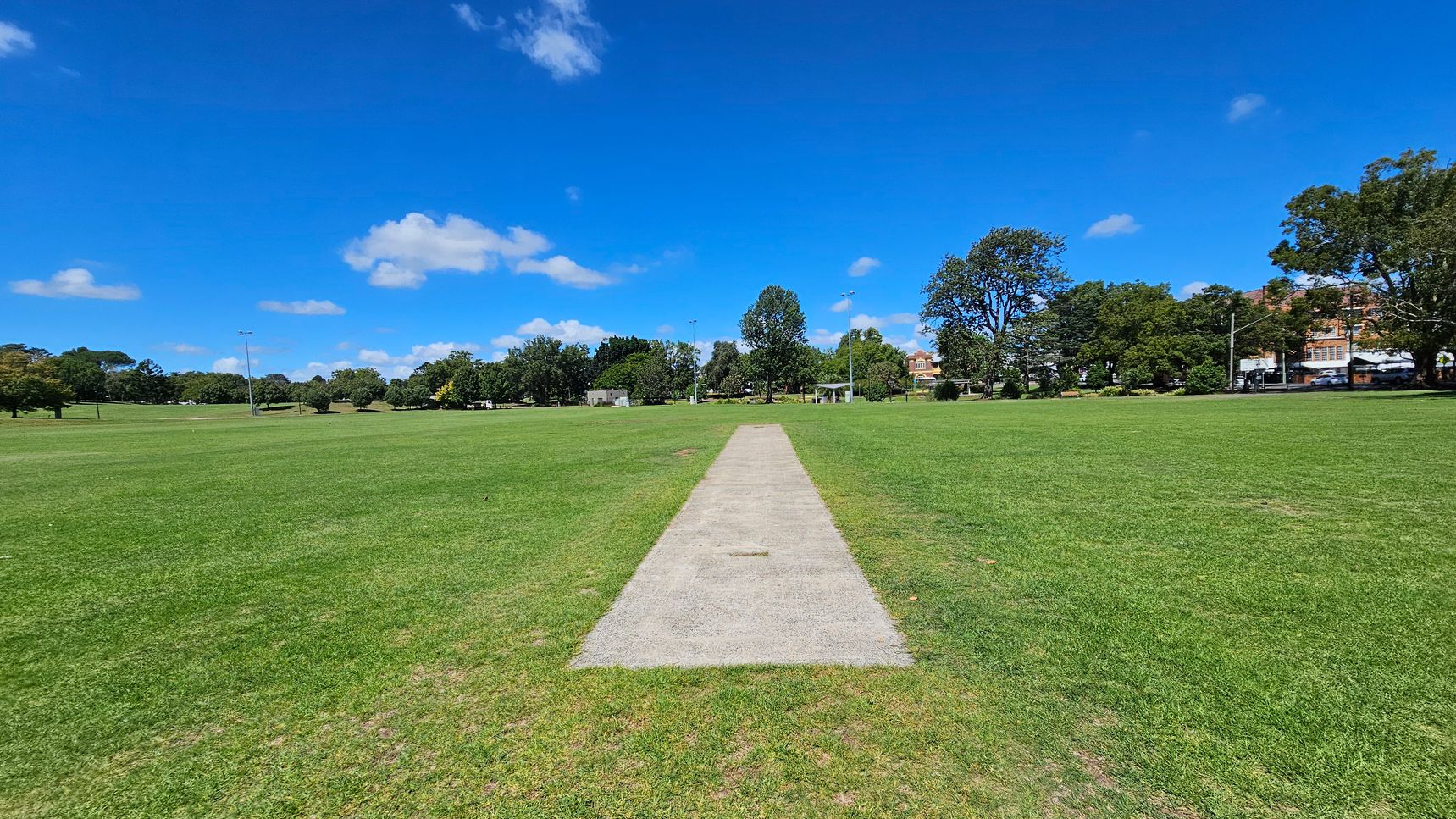A cricket pitch is centred in a large, open grassy field. The pitch is a rectangular strip of lighter-coloured ground. Surrounding the field are trees and some buildings in the distance. There are a few light poles scattered around the field. The sky is clear with a few clouds.