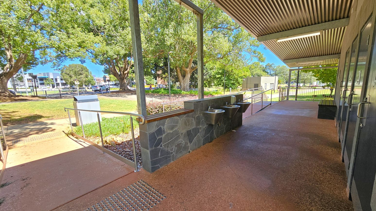 A covered outdoor area with a stone wall featuring two metal sinks on the right. The floor is a textured reddish-brown surface. Metal railings run along a ramp leading to a grassy area with large trees. In the background, there are buildings and a road with cars. The ceiling is corrugated metal with a fluorescent light fixture.