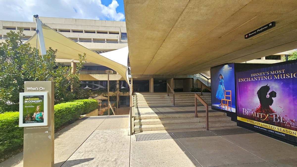 There are steps with handrails on the right, equipped with truncated domes for safety. Large posters and directional signage are positioned on the right wall. On the left, there is a multi-storey building. Additionally, another set of steps on the left, also equipped with truncated domes, leads downward. A concrete facility signage stands on the left. Overhead, a white shade cloth is hanging. Neatly trimmed shrubs border the left side, accompanied by trees.