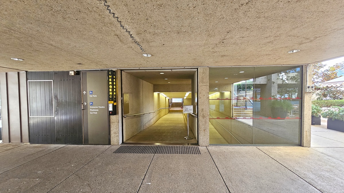 In the middle, there's a ramp with handrails and truncated domes, leading to stairs with handrails. A signage is positioned on the right. Directional signages are posted on the left before the door. The floor and ceiling are concrete. A landscape garden fills the right space.