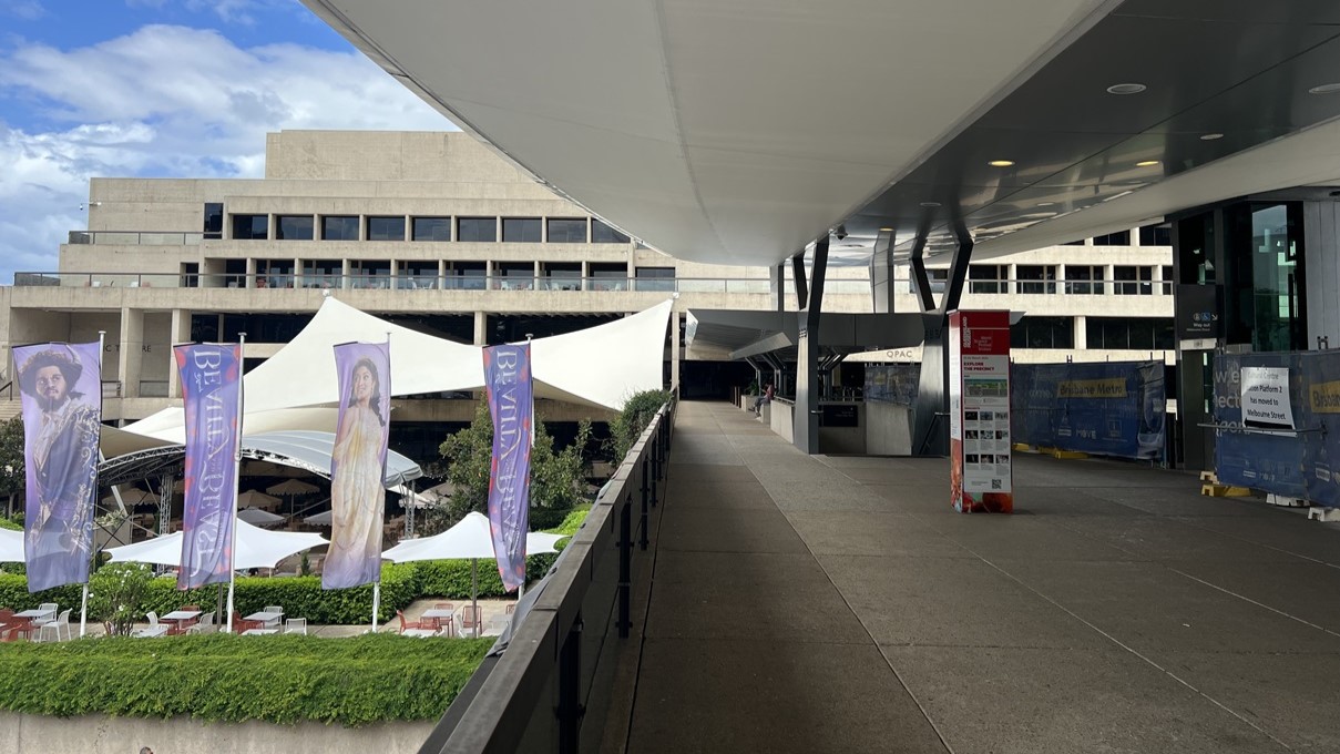 To the left, a row of violet banners line a footpath, with greenery and grass on either side. Tables and chairs are neatly set beneath sizable umbrellas. On the right, a corridor stretches beneath a broad white canopy, guiding towards a multi-storey building. Stairs with handrails descend nearby. Banners and construction barriers are seen on the right side.