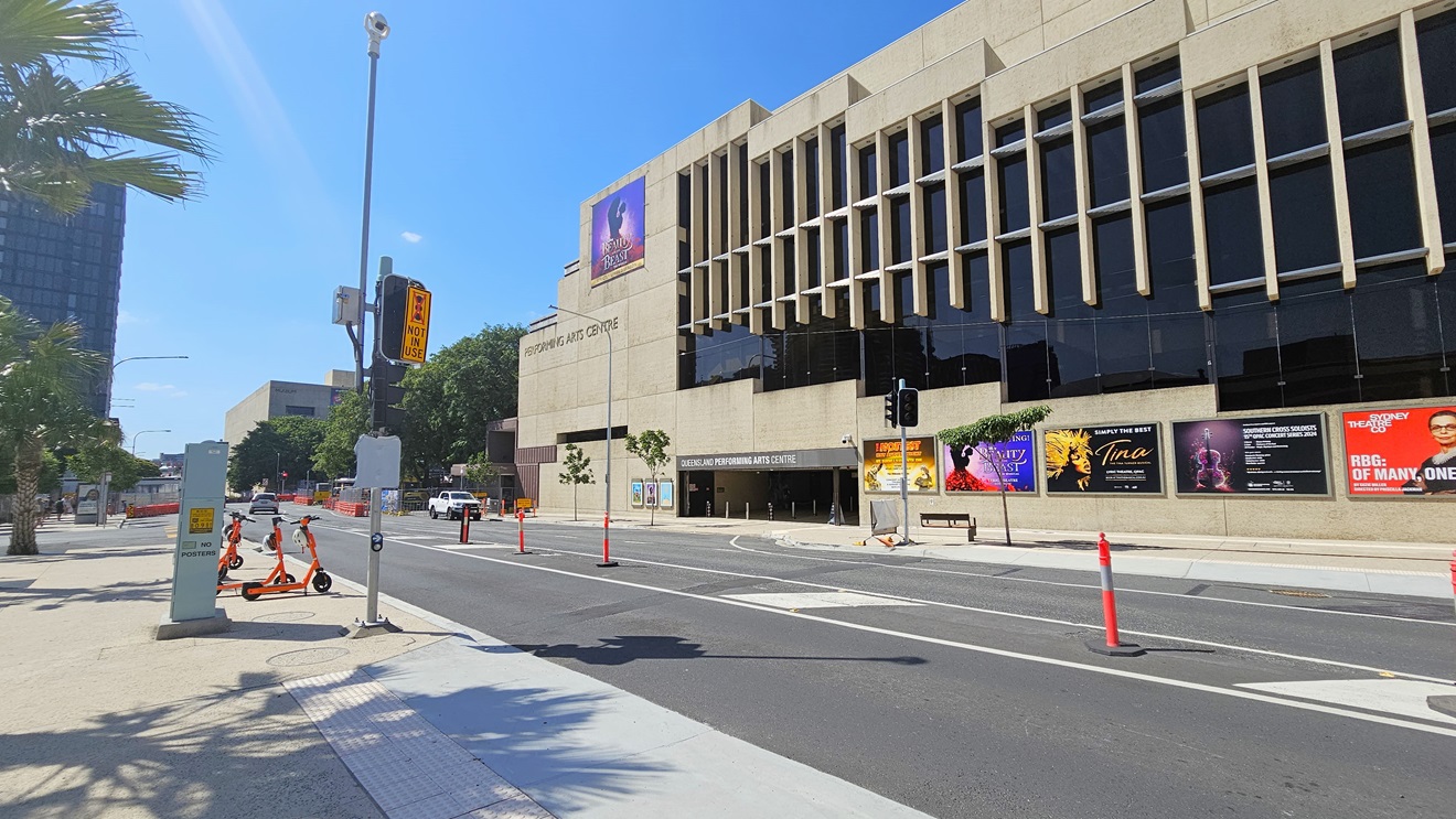 A two-way road features road markings and orange bollards. On the left, a traffic light stands at the road's edge, accompanied by truncated domes, street signs, scooters, trees, and street lights. On the opposite side is a multi-storey building constructed of concrete and glass, displaying the facility's name. Large advertisement posters are affixed to the wall. The building includes an underground car park.
