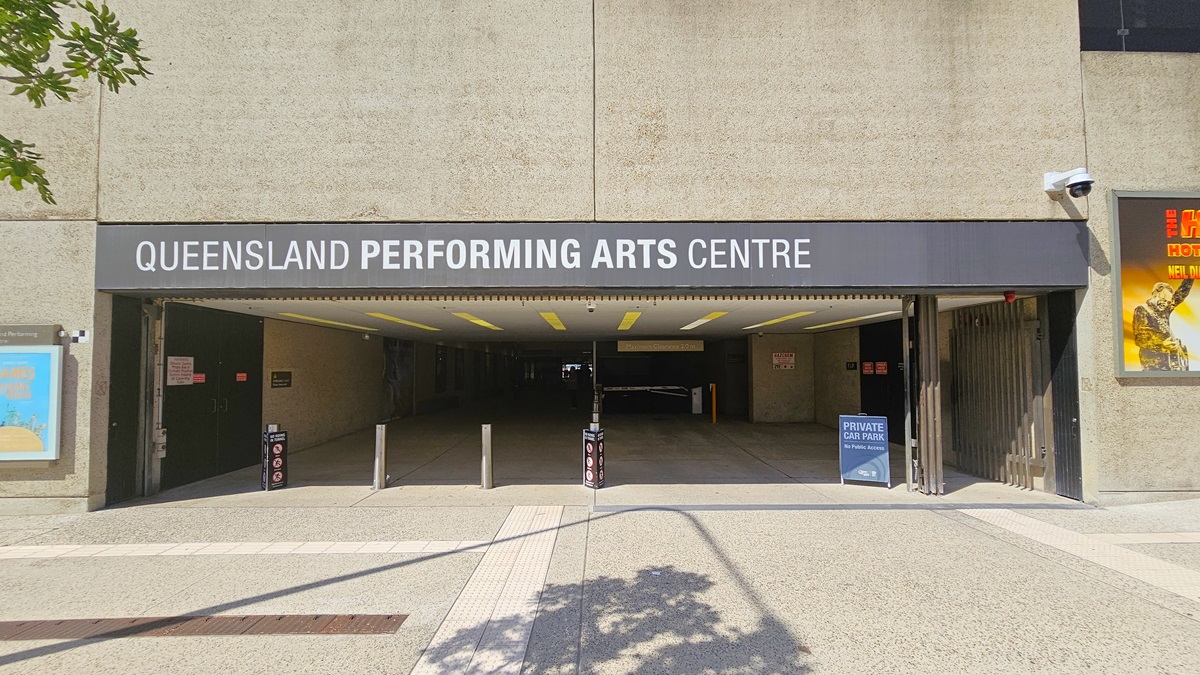 An underground car park, with a low ceiling and dim lighting, has the facility name mounted at the front. Bollards accompanied by warning signs border the left side. A sign stating "Private Car Park" is positioned on the right, with additional signage affixed to the interior walls. Large posters adorn both walls. The building's façade showcases a concrete design, and the floor, featuring truncated domes, has a finished surface.