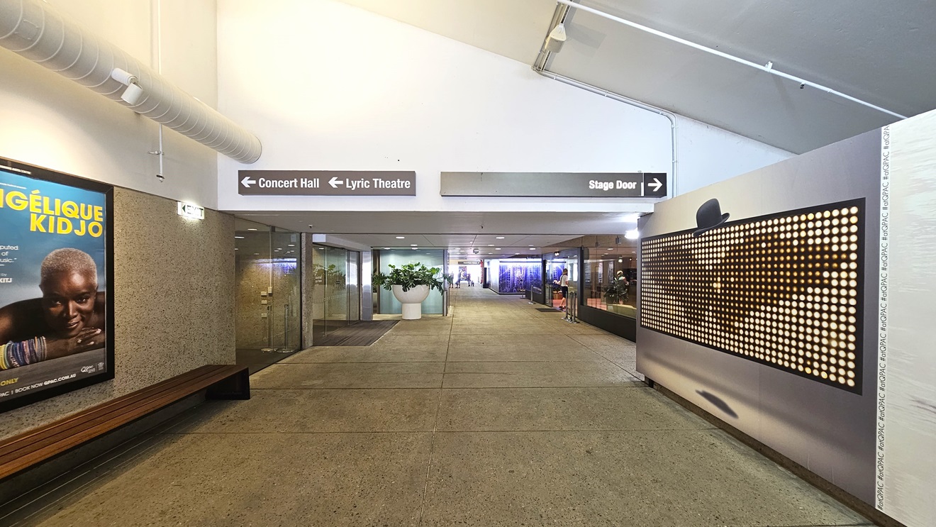 The pedestrian tunnel features a concrete surface and a white, pitched ceiling, fitted with large air conditioning vents and light fixtures. On both walls, large framed posters serve as decorations. To the left, a long bench chair is positioned against the wall. Deeper in the tunnel, shops and directional signs are evident, along with an indoor plant in a sizable pot.