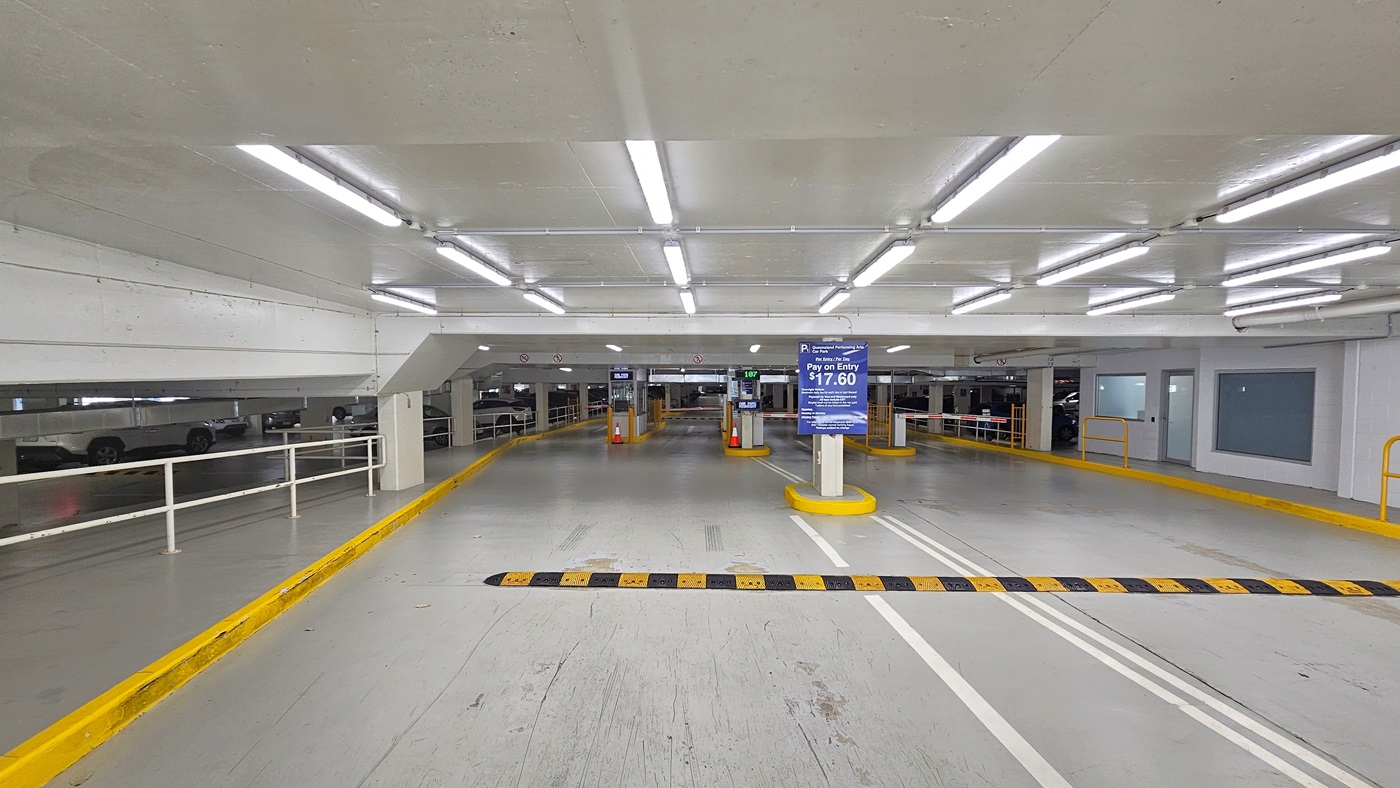 Entrance to the Performing Arts Car Park (P1), featuring a digital sign that shows parking rates on the upper left. To the side, an orange scooter is parked next to a blue parking sign. In the background, stairs ascend to an area lined with trees. Neatly trimmed plants border the left side.