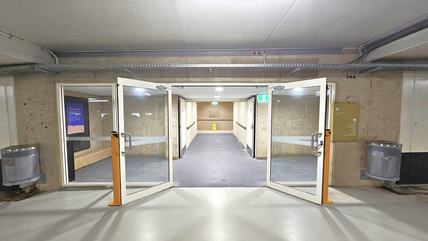 From the underground parking, a manual outward opening glass door leads to stairs and a pedestrian tunnel. Directional signages on both sides, along with a poster on the left wall. A yellow "slippery when wet" sign visible. Concrete floor and white ceiling with stainless structure overhead.