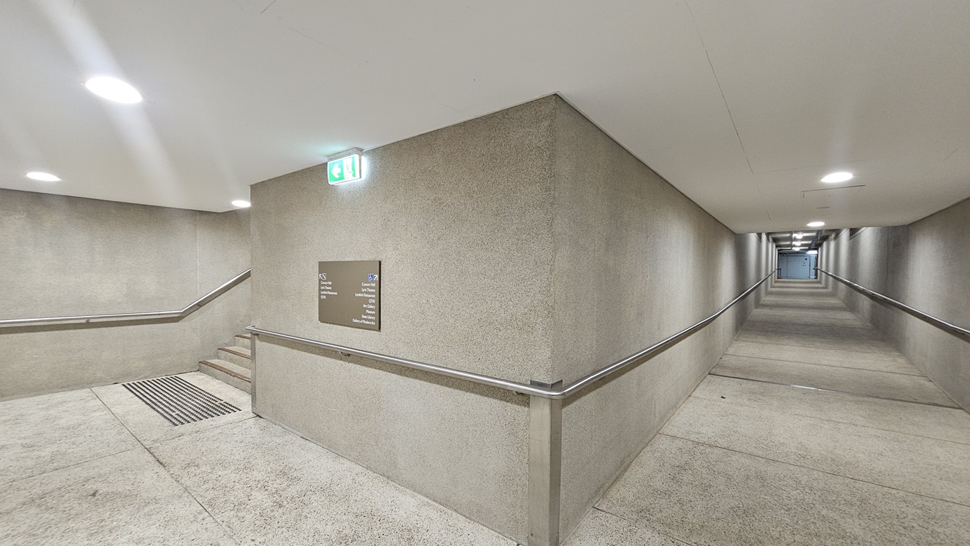 On the right, there is a long pedestrian ramp equipped with handrails. On the left, there are stairs with handrails and truncated domes are installed before the stairs. A directional signage is affixed to the middle wall, along with an exit sign. The walls and floors are made of concrete, and the ceiling is white with LED lights.