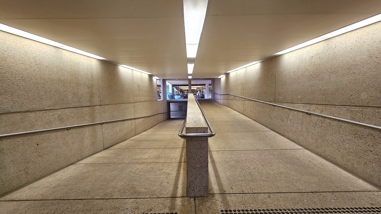 A winding ramp with handrails and truncated domes for safety. A concrete partition secures the ramp. Concrete floor and wall, white ceiling with fluorescent lights. At the end of the ramp, retail shops are visible.
