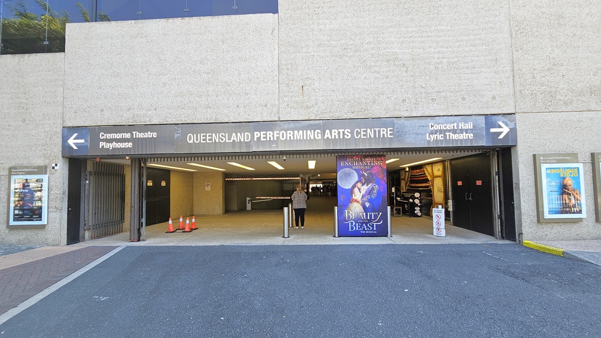 The tunnel entrance with facility signage indicating Queensland Performing Arts Centre and directional arrows to other theatres. Posters decorate the walls. Colourful posters and bollards line the front, alongside orange safety cones and other signs. Inside, retail shops are visible. The building's front has a concrete facade.