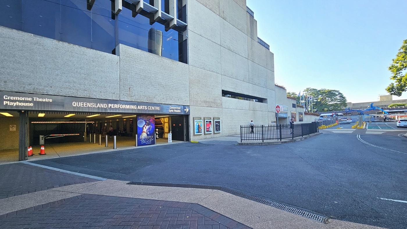 At the tunnel entrance, facility signage directs to Queensland Performing Arts Centre and other theatres. Posters adorn walls. Colourful posters and bollards line the front, with orange safety cones and signs. The building has a concrete and glass facade. To the right, a two-way road with metal fencing and barricaded section for construction.