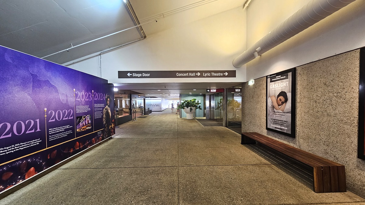 The pedestrian tunnel has a white pitched ceiling with sizable aircon vents on the right. Below, a wooden bench chair. A large poster fills the left wall. Further in, retail shops visible, with indoor plants in a large white pot. Directional signage also present.