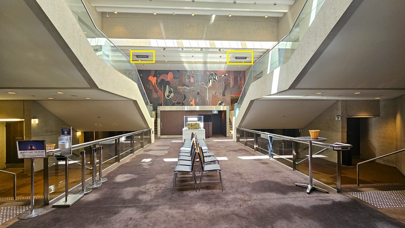 A lobby with beige walls and symmetrical staircases. A large abstract mural at the far end. In the centre, a low table with brochures and movable chairs arranged side by side, flanked by rope bollards. Bright overhead lighting illuminates the space. Directional signages hang from the ceiling on each side of the stairs. Underneath, stairs with handrails and truncated domes are installed on both sides, along with signages. The floor is carpeted.