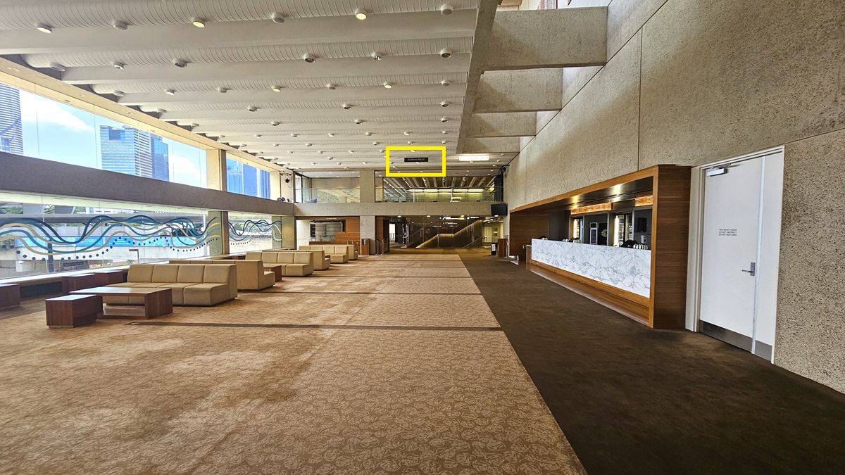 In a spacious lobby with brown printed carpet, a long white marble-fronted bar counter stands to the right. Behind it, fridges stocked with beverages. To the left, lounge chairs and coffee tables are arranged. A directional signage hangs from the ceiling. The wall features white concrete design, while the ceiling is white with large aircon vents and light fixtures. A glass wall with wavy line prints fills the left space.