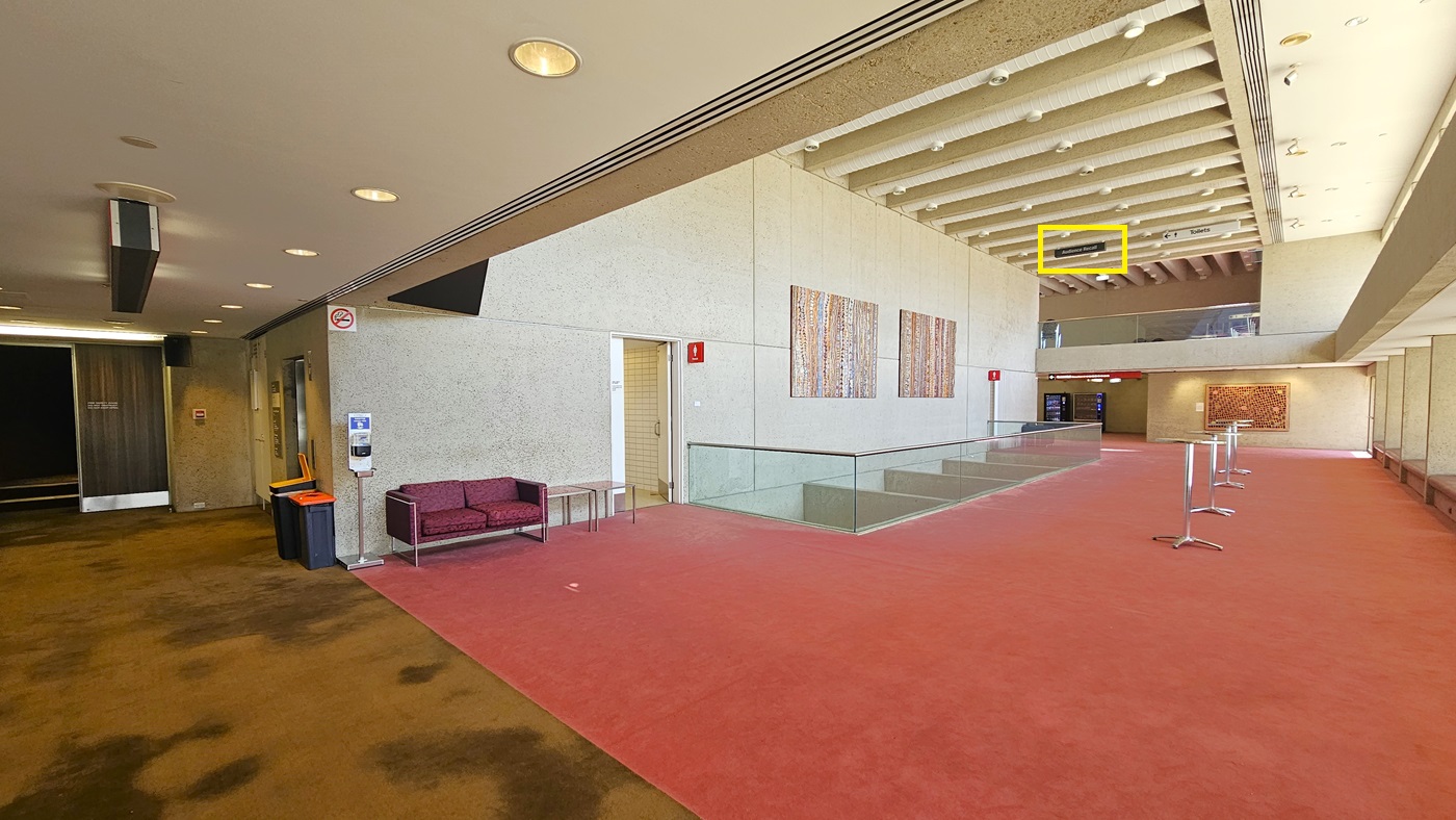 An interior space with a carpeted floor. On the right, a toilet with signage above. Abstract paintings decorate the cream walls. Glass railings enclose the front area, with seating. A cushioned chair and sanitising machine near the toilet door. On the left, steps lead to a darker space. Two rubbish bins nearby. The carpet transitions from pink to brown. The ceiling is white with aircon vents and light fixtures. A directional signage reading "Audience Recall" hangs from the ceiling.