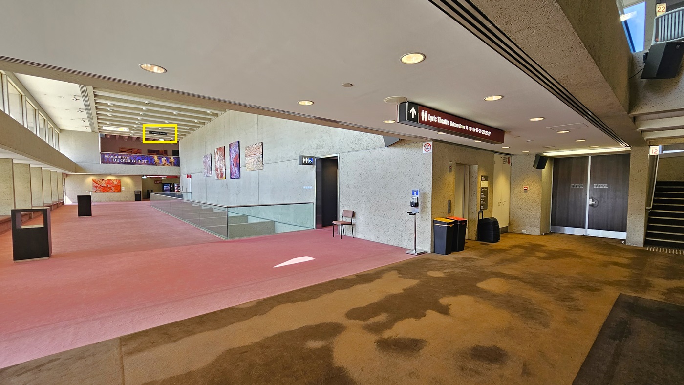 A space with a carpeted floor and a toilet with signage is on the right. Cream walls adorned with abstract paintings. Glass railings enclose the front area, with seating available. A chair and sanitising machine are near the toilet door—steps with handrails on the left lead to the Lyric Theatre balcony. Two rubbish bins and black tables are nearby. The carpet transitions from pink to brown. The ceiling is white with aircon vents and light fixtures. Directional signage hangs from the ceiling.