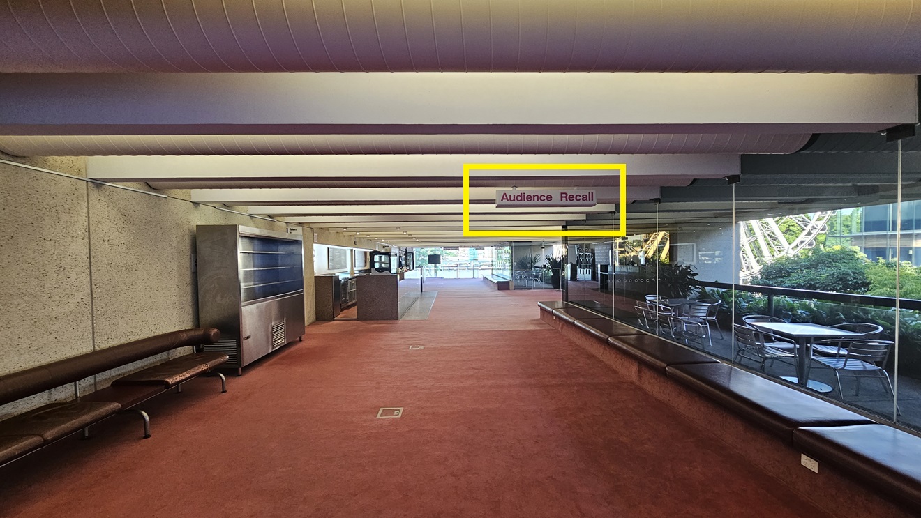 In the carpeted area, cushioned bench chairs are positioned against the walls, accompanied by a stainless steel rack and a reception desk nearby. Tables and chairs are arranged against the wall on the right, with additional seating further inside. The floor is covered in brown carpet, while the white ceiling features large aircon vents and light fixtures. A sign reading "Audience Recall" hangs from the ceiling.