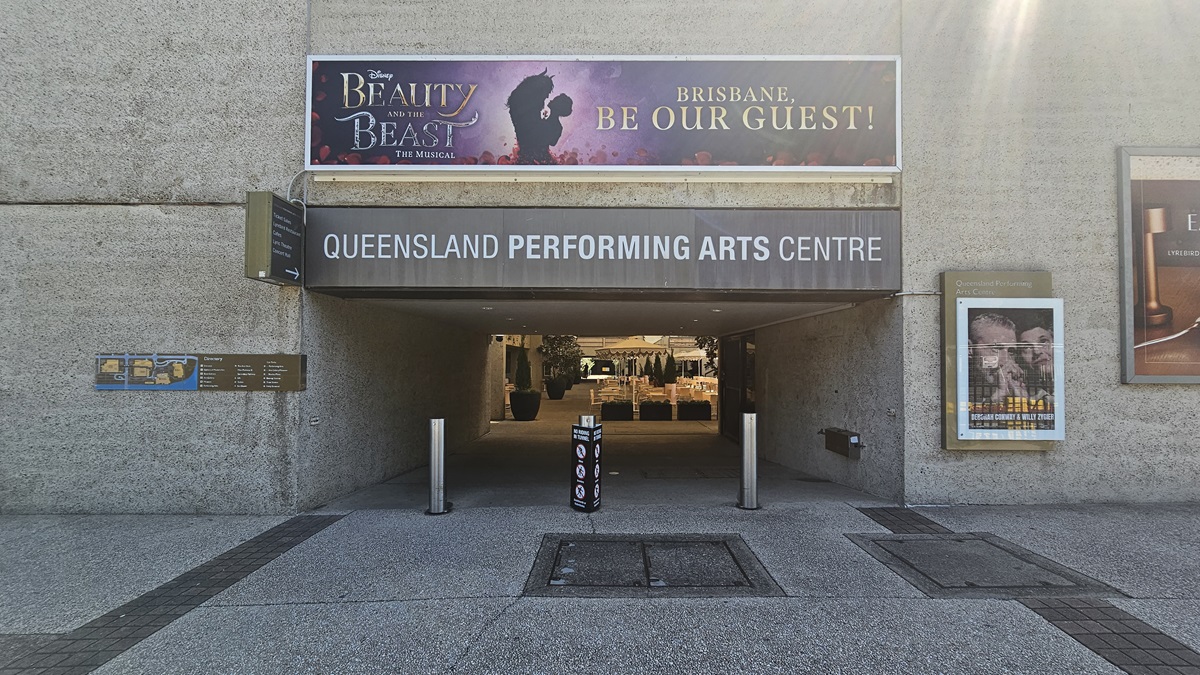 The tunnel features facility signage atop another signage. Additional directional signs and posters adorn the walls. Bollards line the front. Inside, retail shops are visible. The building's front has a concrete facade with black tile flooring.