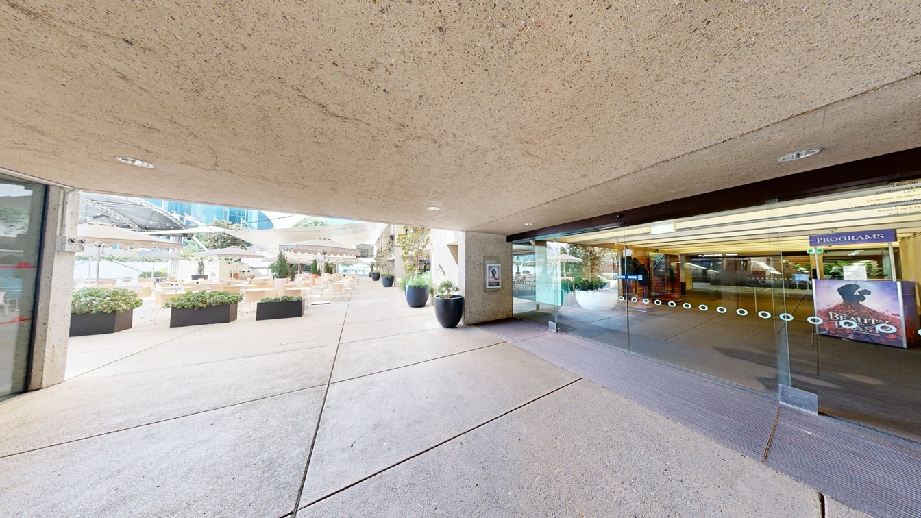 To the right, an automatic glass sliding door equipped with posters. To the left, tables and chairs arranged under large umbrella shades. Large pots of plants on the sides, with plants in planter boxes on the left. The floor and ceiling are concrete.