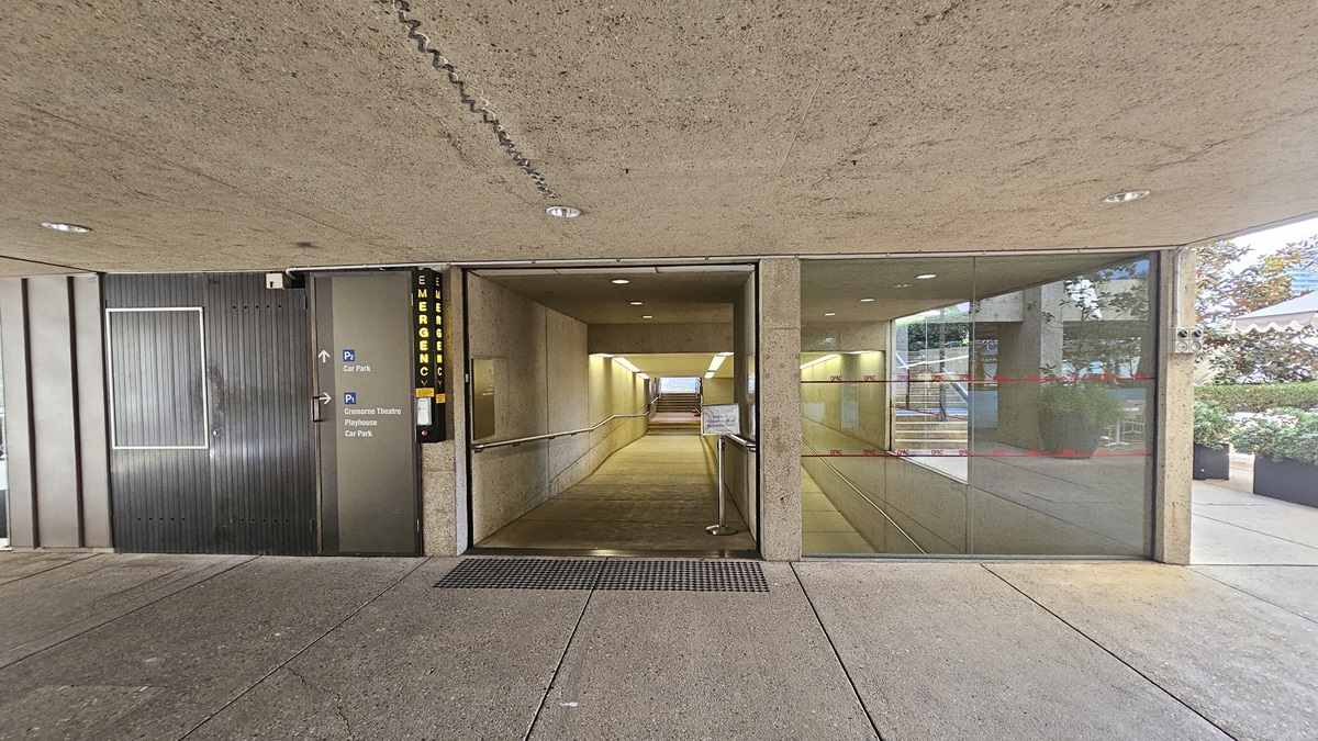 In the middle, there's a ramp with handrails and truncated domes, leading to stairs with handrails. A signage is positioned on the right. Directional signages are posted on the left before the door. The floor and ceiling are concrete. A landscape garden fills the right space.