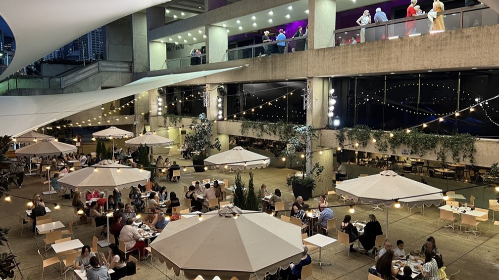 On the left, a spacious outdoor plaza features multiple white umbrellas shading groups of diners at tables, surrounded by trees adorned with string lights. On the right, a layered concrete structure with balconies, each lined with spectators, overlooks the plaza. The floors are tiled, reflecting subtle light, while the ceiling of the overhang is solid concrete with embedded lighting.