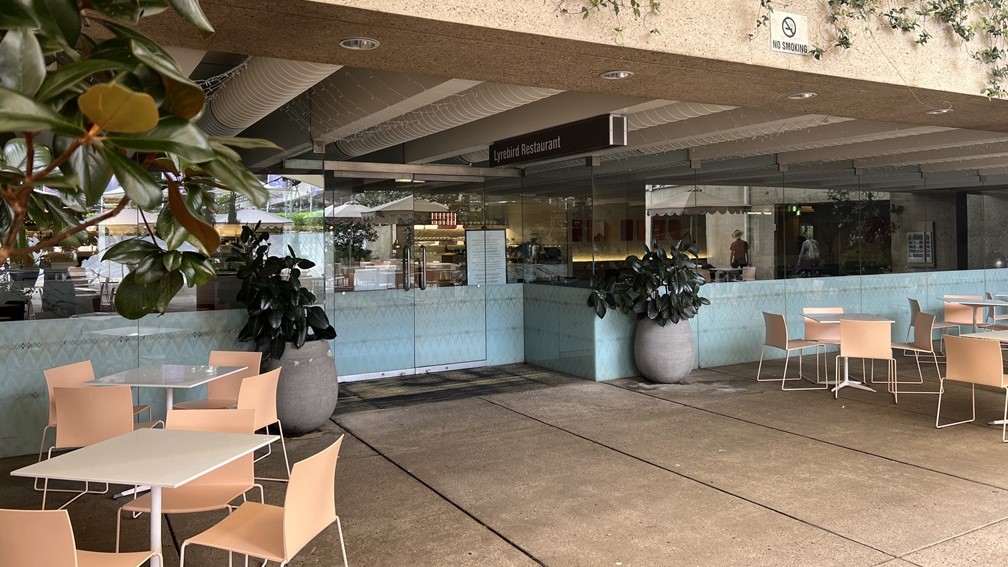 In the foreground, outdoor tables and chairs are set on a grey tiled floor, partly sheltered by a building overhang. Large potted plants line the tiled wall. A glass door leads to "Lyrebird Restaurant", marked by overhead signage. Inside, the decor continues with a similar style, under a roof adorned with white, geometric-patterned panels. The restaurant's ceiling mirrors the outdoor pattern, and pale, reflective tiles cover the interior floor.