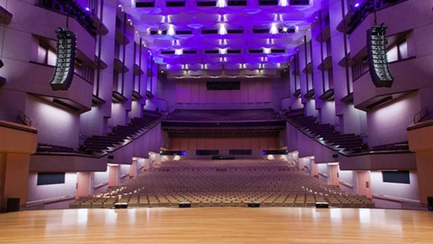 A panoramic view of the concert hall with chairs arranged in rows, stalls, and boxes. Light fixtures hang from the high ceiling. The wall is adorned with timber decorations.