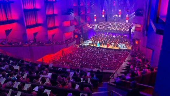 A panoramic view of the concert hall, including the stage, occupied by spectators and performers. The area is dimly lit with lights illuminated from the concert stage. Chairs are arranged in rows, stalls, and boxes. Light fixtures hang from the high ceiling. The wall is adorned with timber decorations.