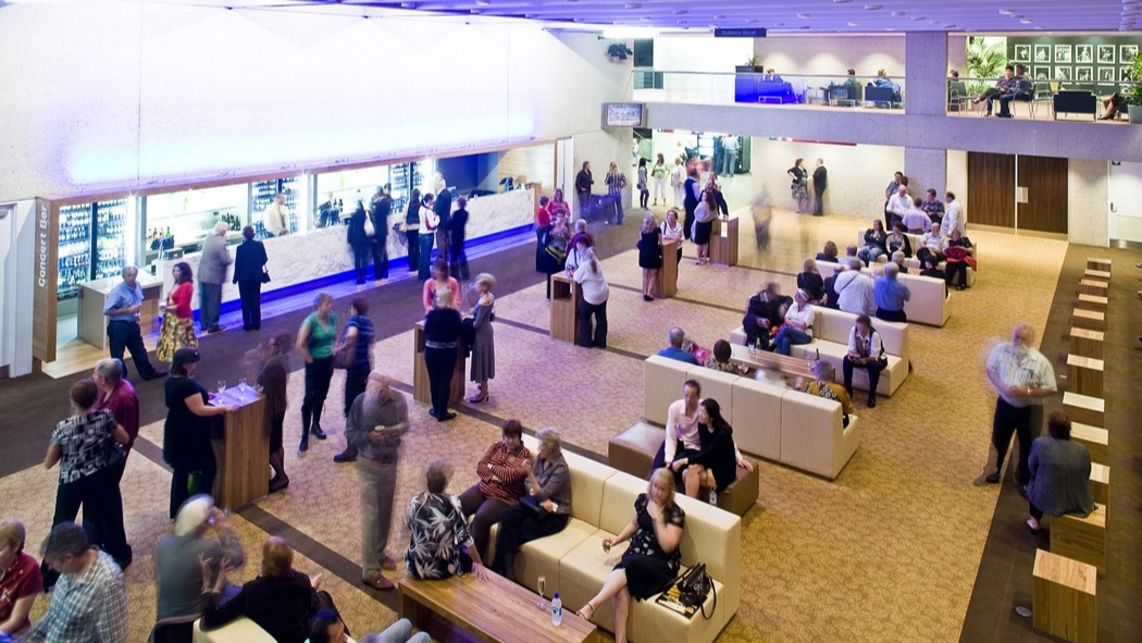 An indoor setting with groups of people mingling. On the left, individuals interact near a bar counter with tall tables and chairs. On the right, more people gather, some seated on sofas. The mezzanine features occupied tables and chairs. Varied seating arrangements fill the foreground. The carpeted floor boasts a pattern, while embedded lights illuminate the ceiling.
