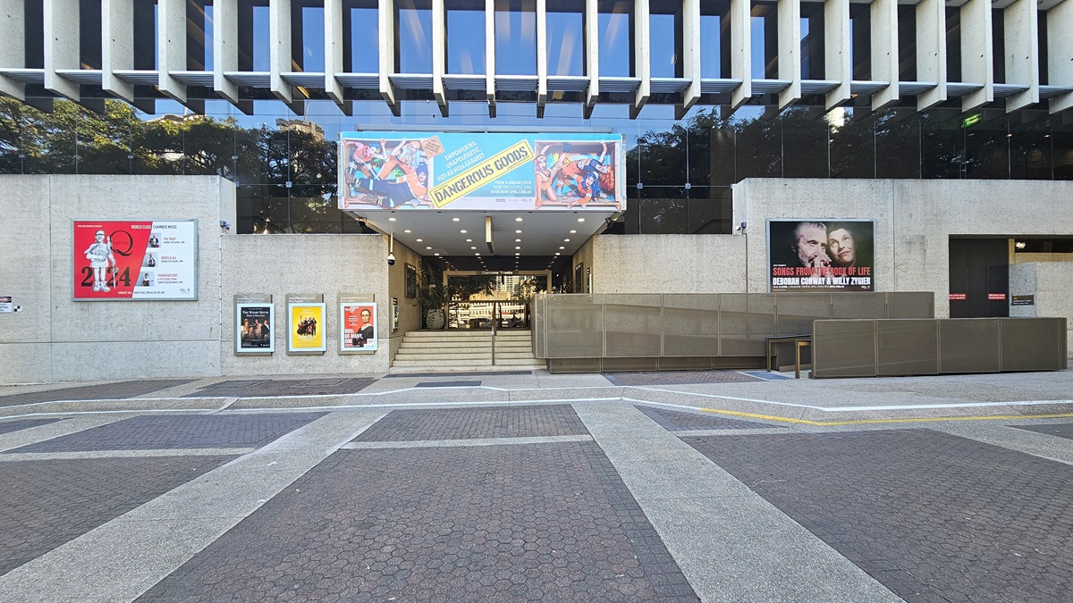 To the left, a pedestrian pathway is lined with large trees. On the right, a building with a textured concrete exterior is adorned with advertisement posters. In the centre foreground, three metal bollards stand evenly spaced across the pathway. The walkway extends towards the end of the building.