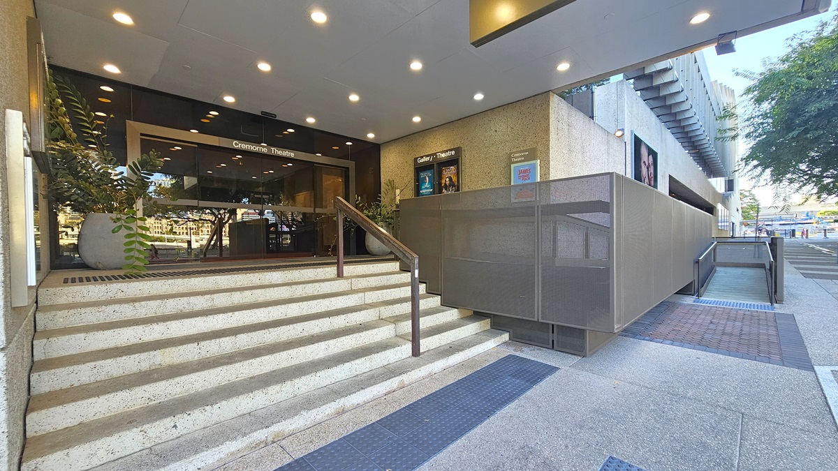 Entrance to Cremone Theatre. Steps with metal handrails and truncated domes lead to the left, while a ramp with handrails, also equipped with truncated domes, is on the right. The building's entrance features an automatic glass sliding door, with large potted indoor plants decorating the front. Posters are affixed to the walls on the right. The ceiling is white with LED lights.