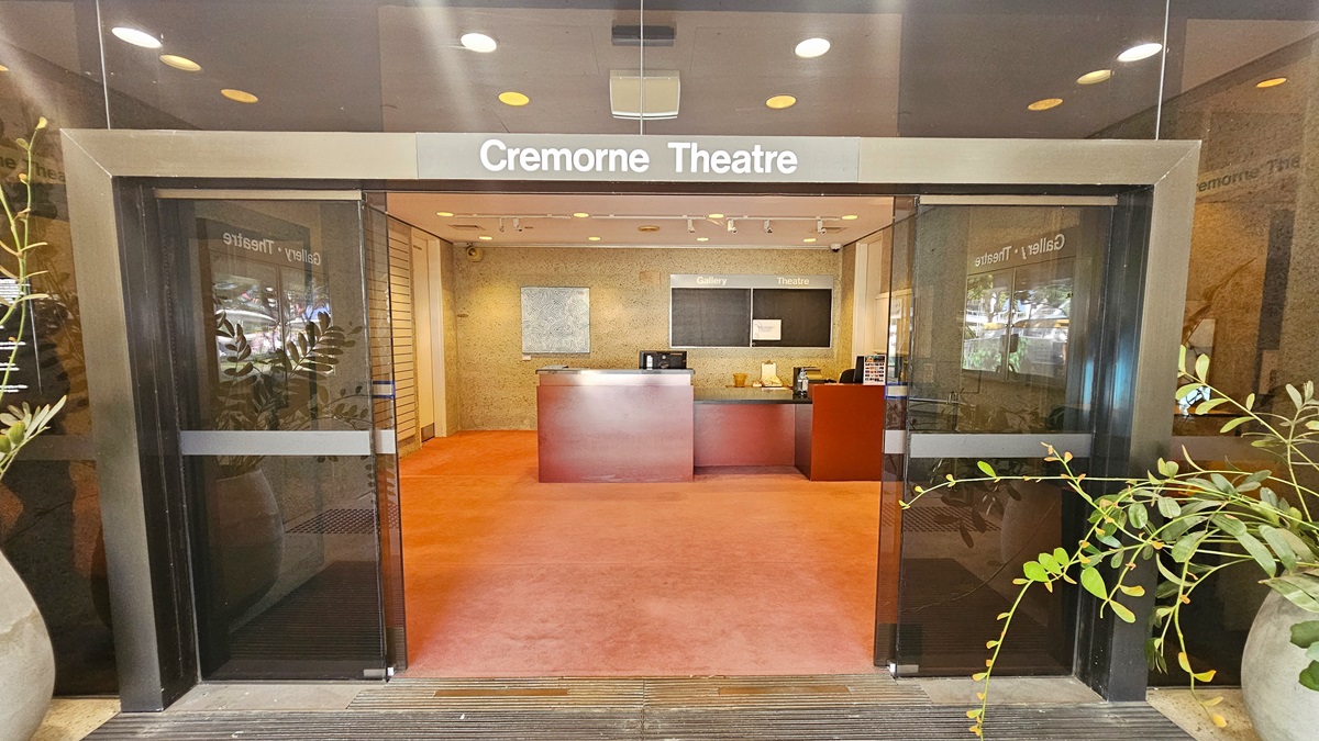 The entrance features an automatic glass sliding door. a signage that reads  "Cremorne Theatre" is placed above. Inside, a reception desk with a brown facade holds computers and posters. Behind the desk, an abstract painting hangs on the cream wall. The floor is covered in pink carpet, and the ceiling features white light fixtures. Large potted plants adorn the entrance.