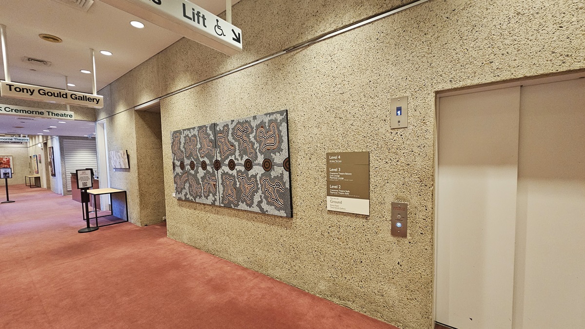 In the lobby, a pink carpet covers the floor. Tables and signage are positioned against the cream wall, which also displays abstract paintings. Directional signage hangs from the ceiling. To the right, there's a lift accompanied by directional signage and a large abstract painting. The ceiling is white with LED lights.