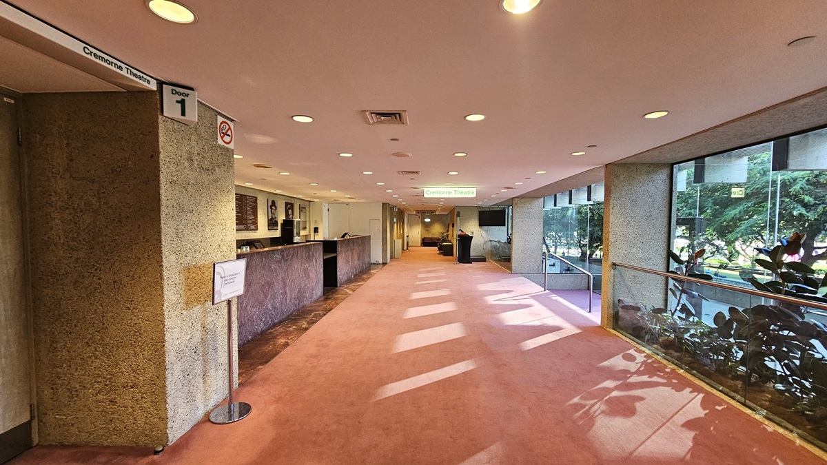 In the lobby, a pink carpet covers the floor. A marble-fronted reception desk is situated on the left, adorned with photos and signage on the wall behind it. To the right, there are stairs with handrails and glass walls. Ornamental plants and large trees border the right side. The ceiling is white with LED lights.