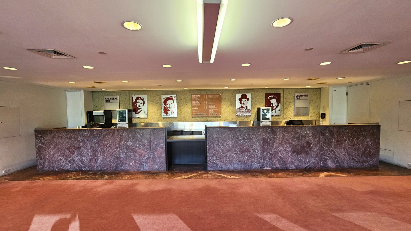 The lobby presents a lengthy front desk with a marble façade, adorned with framed photos and posters against a beige wall. Computers and additional posters rest on the counter. Pink carpeting lines the floor, while the ceiling is white with both LED and fluorescent lights, along with air conditioning.
