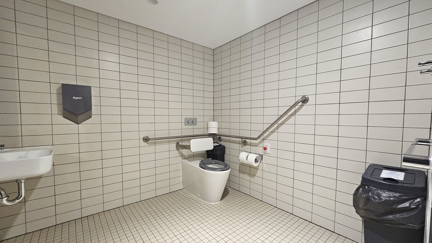 The accessible bathroom includes a toilet bowl with a corner hand grab, alongside a tissue dispenser, a rubbish bin, and a red push button for locking. A wash basin and additional tissue dispenser are mounted on the left wall. The floor is tiled with small white tiles, while the wall features larger white tiles, and the ceiling is white.