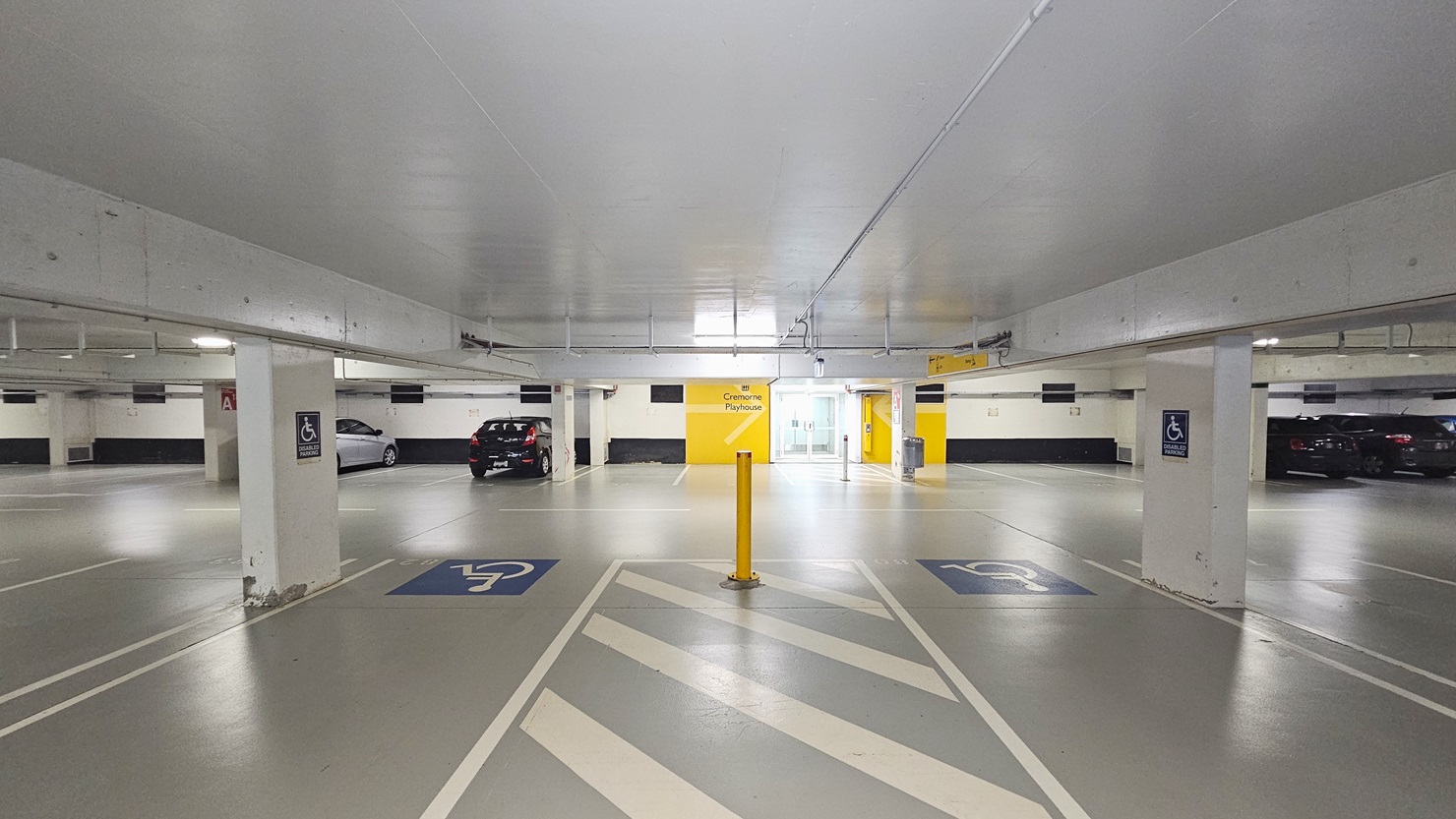 The accessible parking area is marked with designated accessibility symbols, accompanied by a yellow bollard in the centre. The ceiling is low and painted white, with a visible glass door entrance at the far end. Yellow directional signage is positioned on the middle wall, while vehicles are parked on the opposite side.