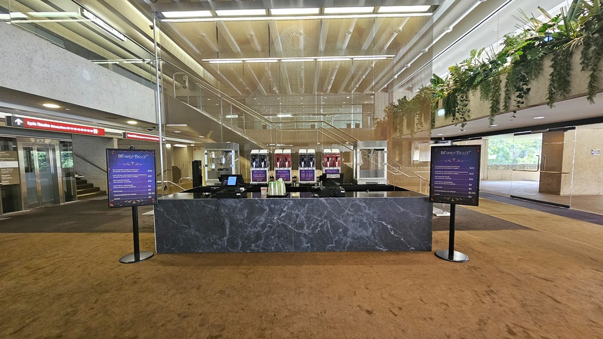 In the middle, there is a lift equipped with a poster. Stairs with handrails and truncated domes are installed on both sides of the lift. Another set of descending stairs with handrails and truncated domes is also installed on both sides of the stairs. The ceiling and wall are made of concrete and have air conditioning vents.