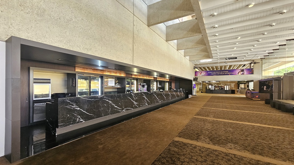 The lobby showcases a long front desk with a marble façade and embedded monitors against a beige wall. The carpeted floor extends towards the background, leading to a seating area with sofas. Various drinks are displayed in the fridge. The ceiling is lined with parallel lighting fixtures and aircon vents.
