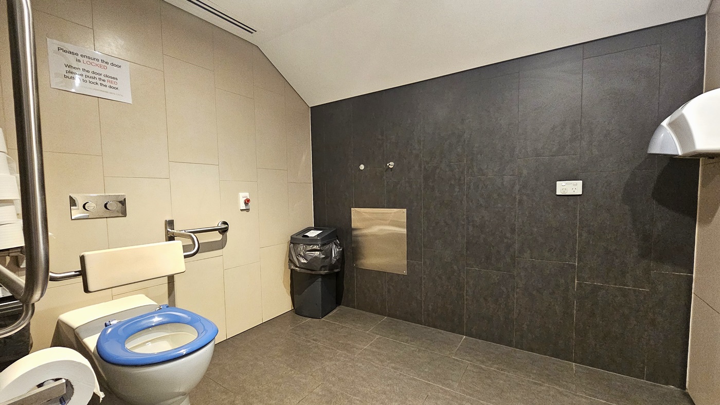 The accessible bathroom has a toilet bowl with a hand grab positioned on the left, along with a tissue dispenser and rubbish bin. Another rubbish bin is set against the wall on the right. A reminder is affixed to the upper wall, and a red push button to lock the door is also visible. The floor has grey tiles, and the ceiling is white. The wall combines black and cream tiles. A hand dryer is positioned on the right.