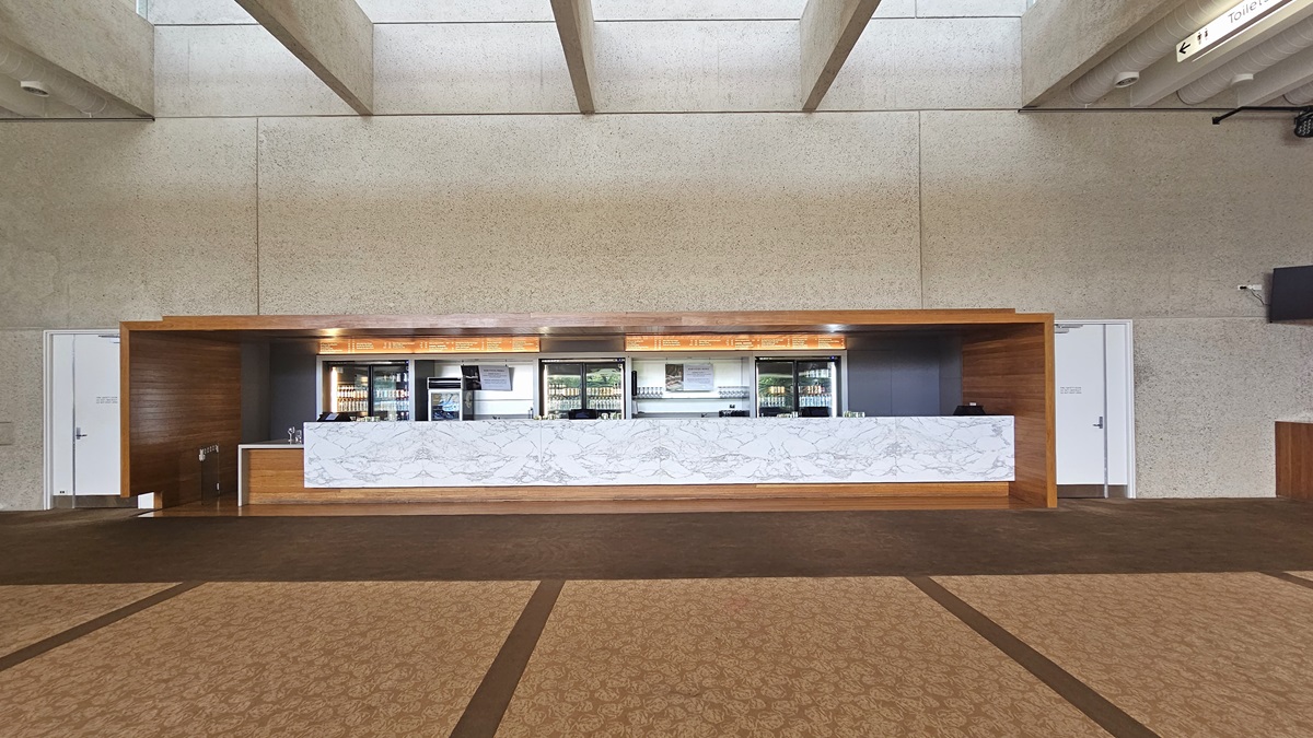 The lobby showcases a long front desk with a wooden and marble façade against a beige wall. Various drinks are displayed in the fridge. Adjacent is a white door. The floor has printed brown carpet, and the ceiling is lined with parallel lighting fixtures and aircon vents.