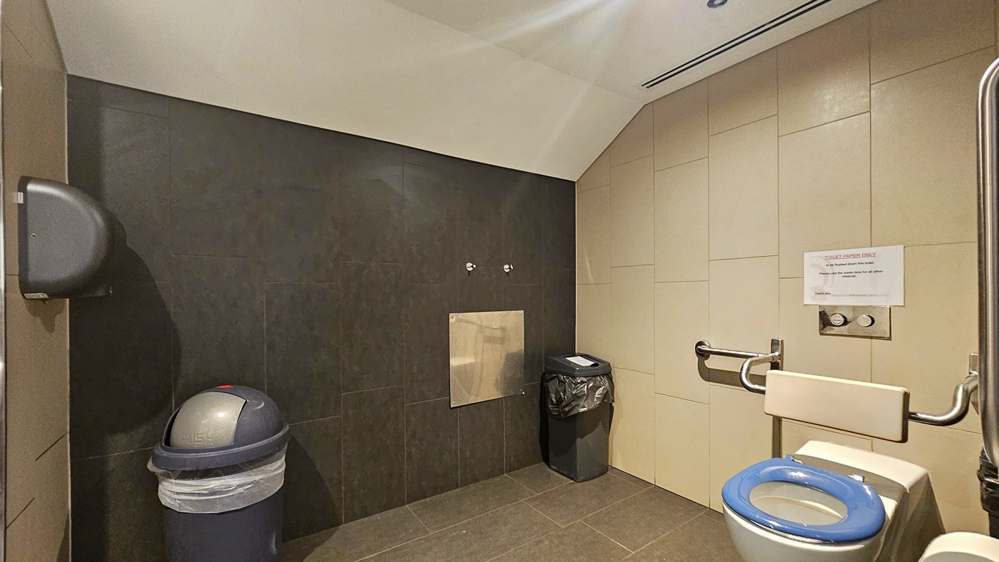 The accessible bathroom has a toilet bowl with a hand grab positioned on the right, along with a tissue dispenser and rubbish bin. Another rubbish bin is set against the wall on the left. A reminder is affixed to the wall. The floor has grey tiles, and the ceiling is white. The wall combines black and cream tiles. A hand dryer is positioned on the right.