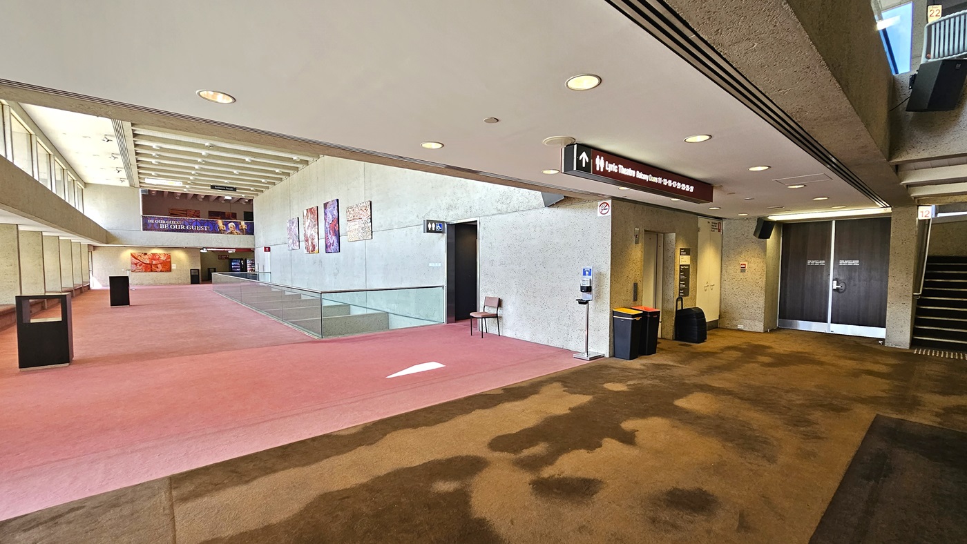 An interior space with a carpeted floor; on the left, there's an accessible toilet with signage above. Posters are affixed to the cream walls. The front is enclosed with glass railings with seating. A chair is positioned near the toilet door along with a sanitising machine. On the right, a staircase with railings leading up, a lift, and a rubbish bin nearby. The carpet transitions from pink to brown and the ceiling is white with LED lights.