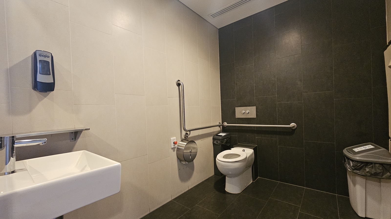 The accessible bathroom features a toilet bowl with a hand grab positioned on the corner, along with a tissue dispenser and rubbish bin. Another rubbish bin is set against the wall on the right. A reminder is affixed to the wall, and a red push button to lock the door is also visible. A wash basin is mounted to the wall on the left along with a liquid soap dispenser. The floor has grey tiles, and the ceiling is white. The wall combines black and cream tiles.