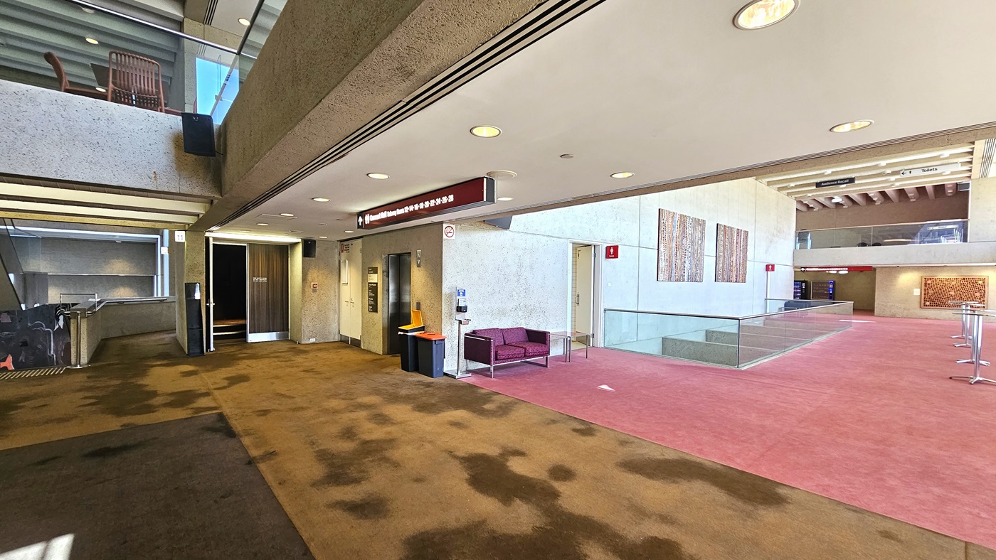 An interior space with a carpeted floor; on the right, there's  toilet with signage above. Posters are affixed to the cream walls. The front is enclosed with glass railings with seating. A cushioned chair is positioned near the toilet door along with a sanitising machine. On the left, a ramp with railings leading up, a lift, and a rubbish bin nearby. The carpet transitions from pink to brown and the ceiling is white with LED lights.