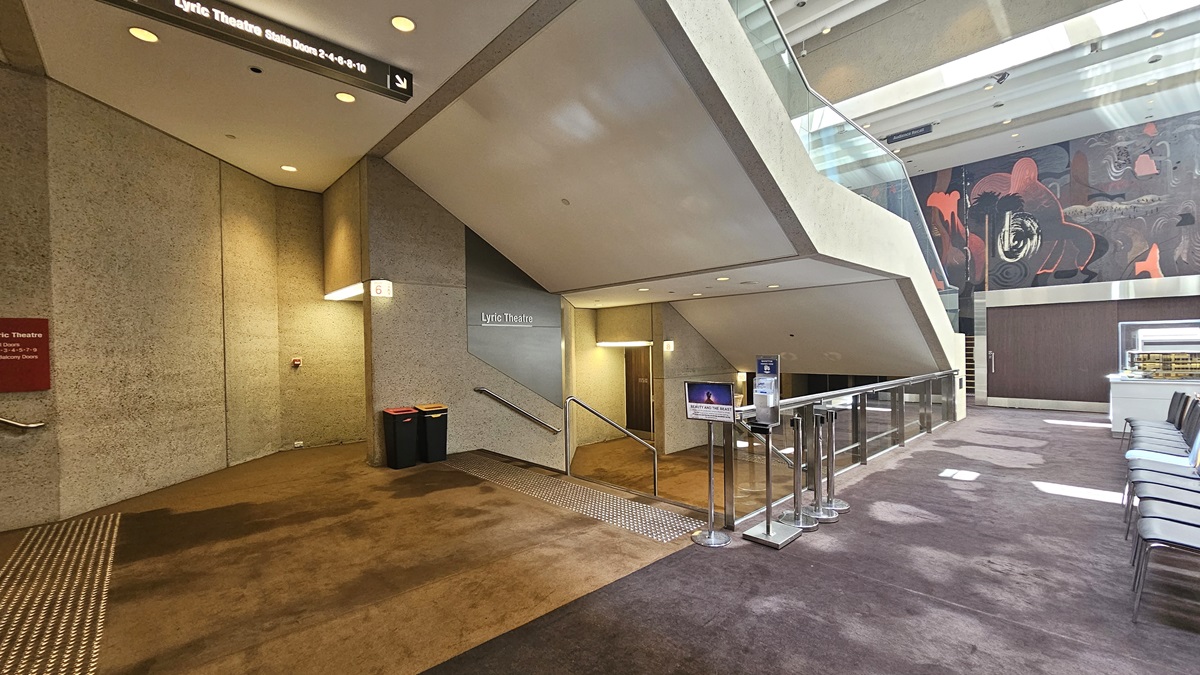 A spacious lobby with beige walls and symmetrical staircases on either side. A large abstract mural at the far end. In the centre, a low table with brochures, and movable chairs arranged side by side, flanked by rope bollards. Bright overhead lighting illuminates. Stairs with handrails and truncated domes are installed on both sides, along with signages. The floor is carpeted.