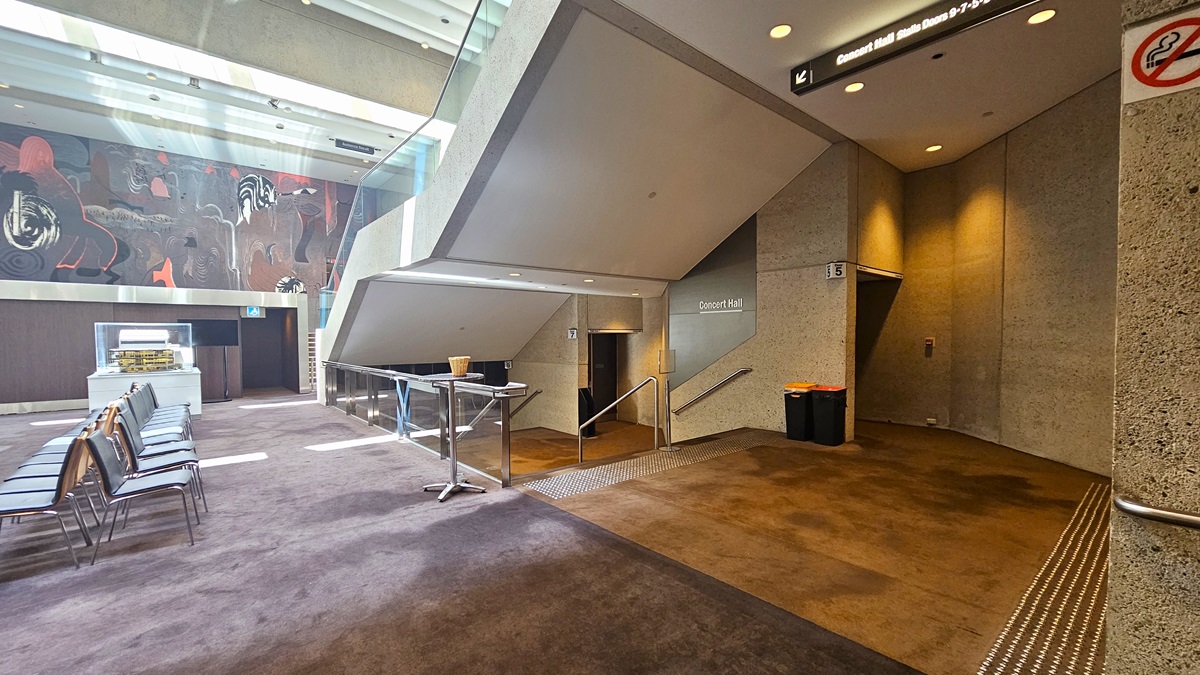 To the right, there is a staircase with handrails equipped with truncated domes, while a large mural decorates the wall. Underneath, there is another set of staircases with truncated domes and handrails leading to the Concert Hall. Signages are affixed to the walls, and two rubbish bins are placed beside them. The floor is carpeted, and the ceiling is white.