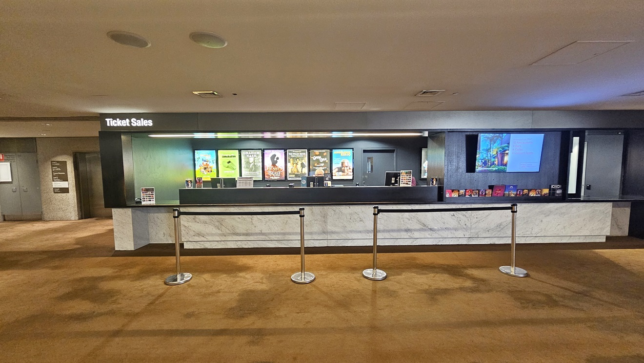 The ticket sales office combines black and marble design. A flatscreen TV is mounted on the left side, with large advert posters on the wall. Brochures are displayed below the TV, and a queue bollard is positioned in front. Directional signages are visible on the left. The floor is carpeted, and the ceiling is white with LED lights and air conditioning.