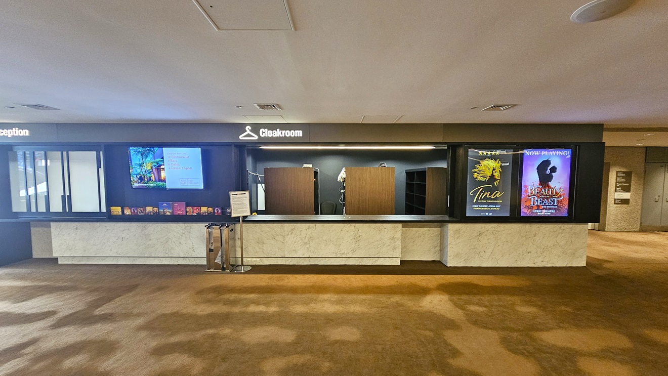 The reception and cloakroom feature a black and marble design. To the left is the reception area with a flatscreen TV mounted on the wall and brochures displayed below. A dispenser for plastic for wet umbrellas and a reminder signage are also present. On the right is the cloakroom with cabinet racks in a wooden design. Large advert posters are affixed to the outer wall. The floor is carpeted, and the ceiling is white with LED lights and air conditioning.