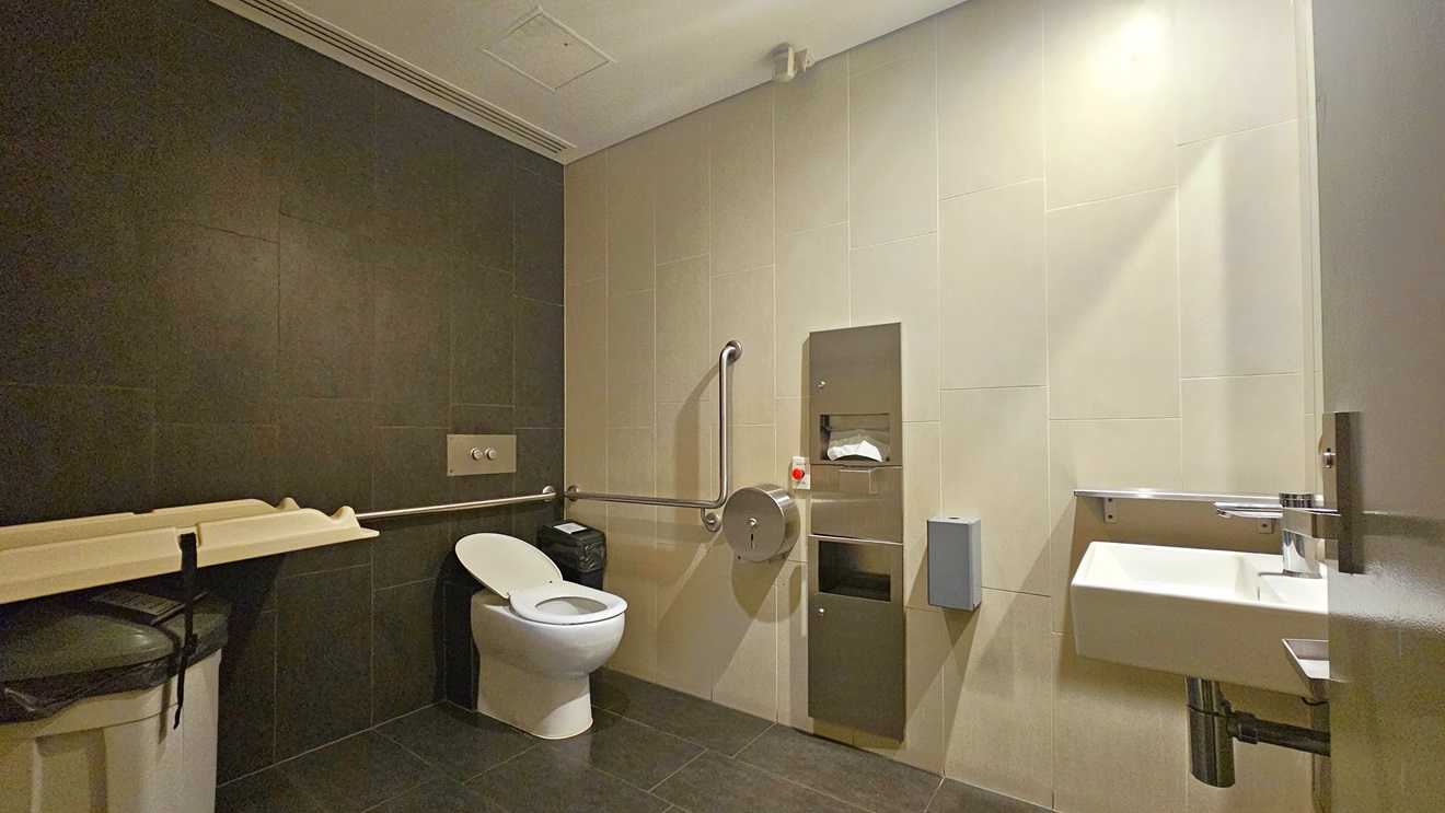 The accessible bathroom features a toilet bowl with hand grabs on the corner and tissue dispensers mounted to the wall. A change table is positioned to the left, with a rubbish bin underneath. To the right is a wash basin. The wall has black and white tiles, and the floor is black.